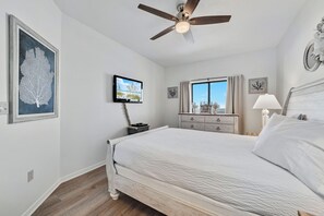 Une chambre à coucher comprenant un lit, un ventilateur de plafond, une télévision, une commode et une fenêtre avec des rideaux.
