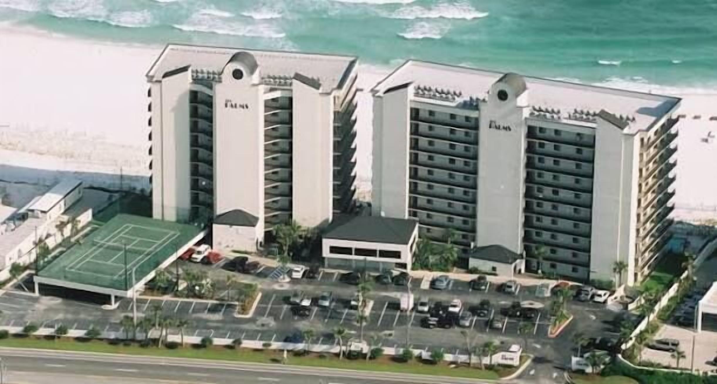 A multi-story hotel with a central courtyard, parking lot, and a beach view.