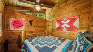 Ein gemütliches Cabin-Zimmer mit Holzvertäfelung, einem Deckenventilator, einem Bett mit einem gemusterten Quilt und zwei Wandbildern.