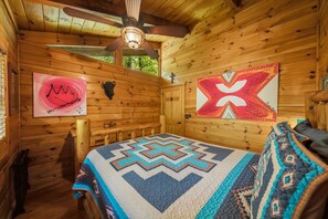 A cozy cabin room with a wooden interior, a ceiling fan, a bed with a patterned quilt, and two wall decorations. - Marigold's Mountain Memories (Gatlinburg)