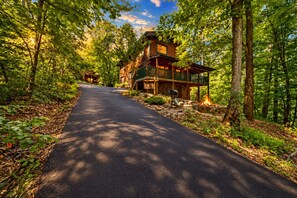 Une cabane en bois, construite sur des fondations en pierre, entourée d’une zone boisée et accessible par une allée pavée menant à l’entrée.