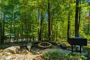 A forested area with a stone fire pit surrounded by rocks and a black sign. - Marigold's Mountain Memories (Gatlinburg)