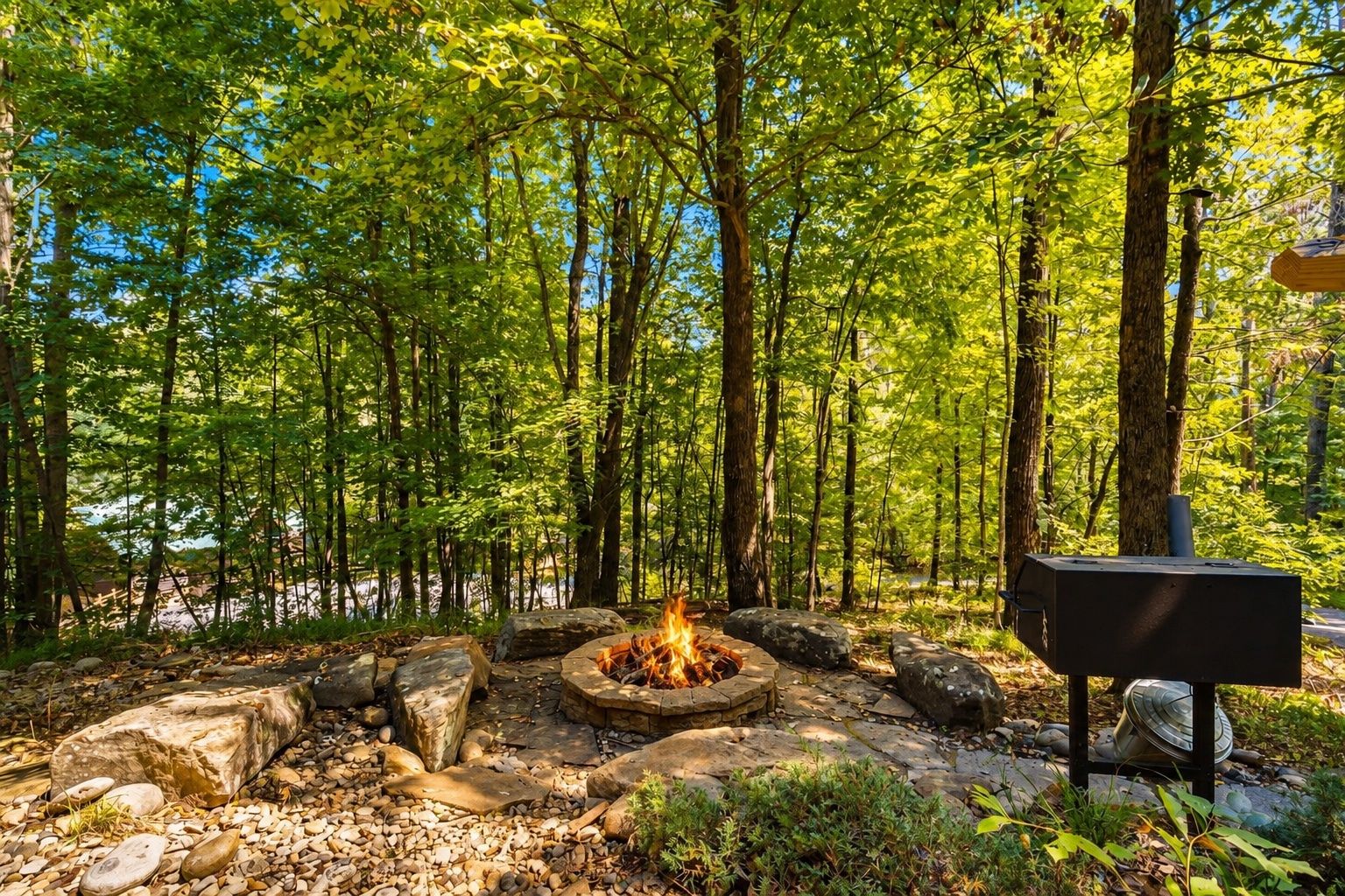 A forested area with a fire pit surrounded by rocks and a grill.