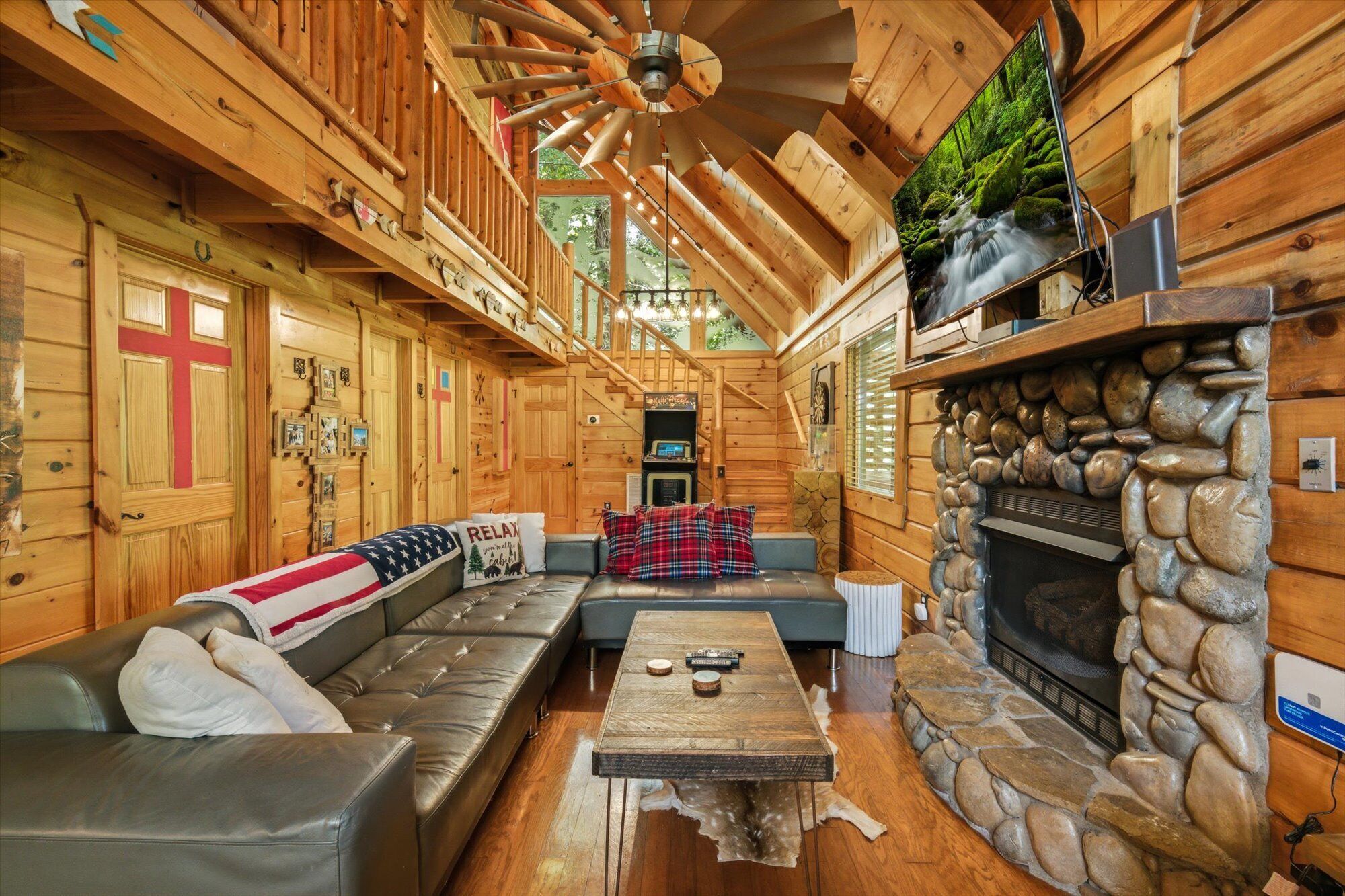 A log cabin interior with a stone fireplace, leather sofa, and a wooden ceiling fan.