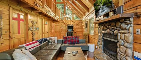 A log cabin interior with a stone fireplace, leather sofa, and a wooden ceiling fan.