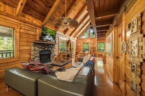 A log cabin interior with a stone fireplace, leather sofa, wooden dining table, and a ceiling fan. - Marigold's Mountain Memories (Gatlinburg)