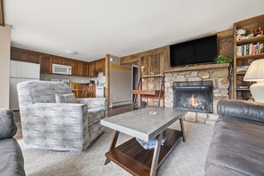 A cozy living room with a fireplace, a television, and a kitchen area with wooden cabinets. - Three Bedroom condo- 200 yard walk to ski slopes (Sugar Mountain)