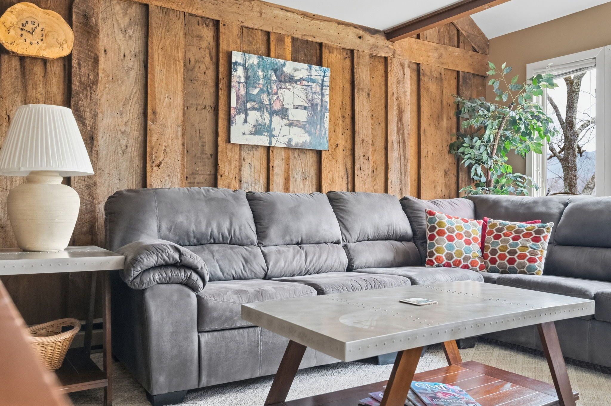 A living room with a grey sofa, a wooden wall, a painting, and a potted plant.