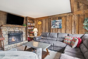Ein gemütliches Wohnzimmer mit Kamin aus Stein, einem grauen Sofa und einem flachbildfernseher an der Wand.