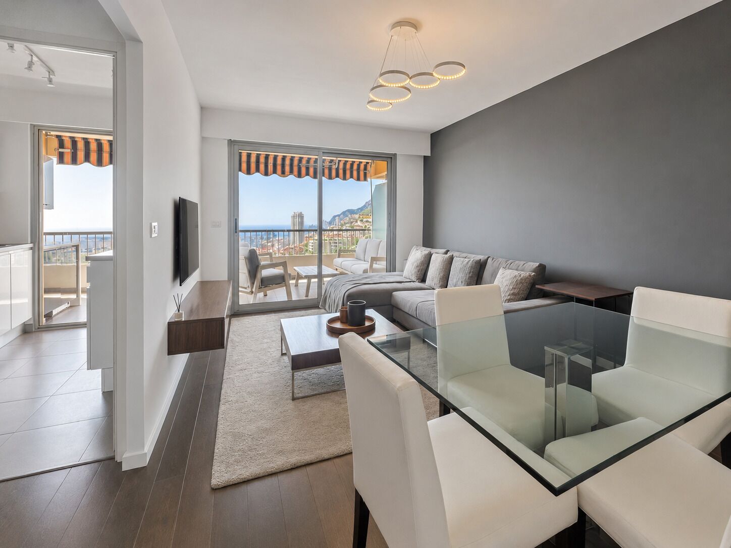 A modern living room with a glass dining table, a sofa, and a balcony with a view.