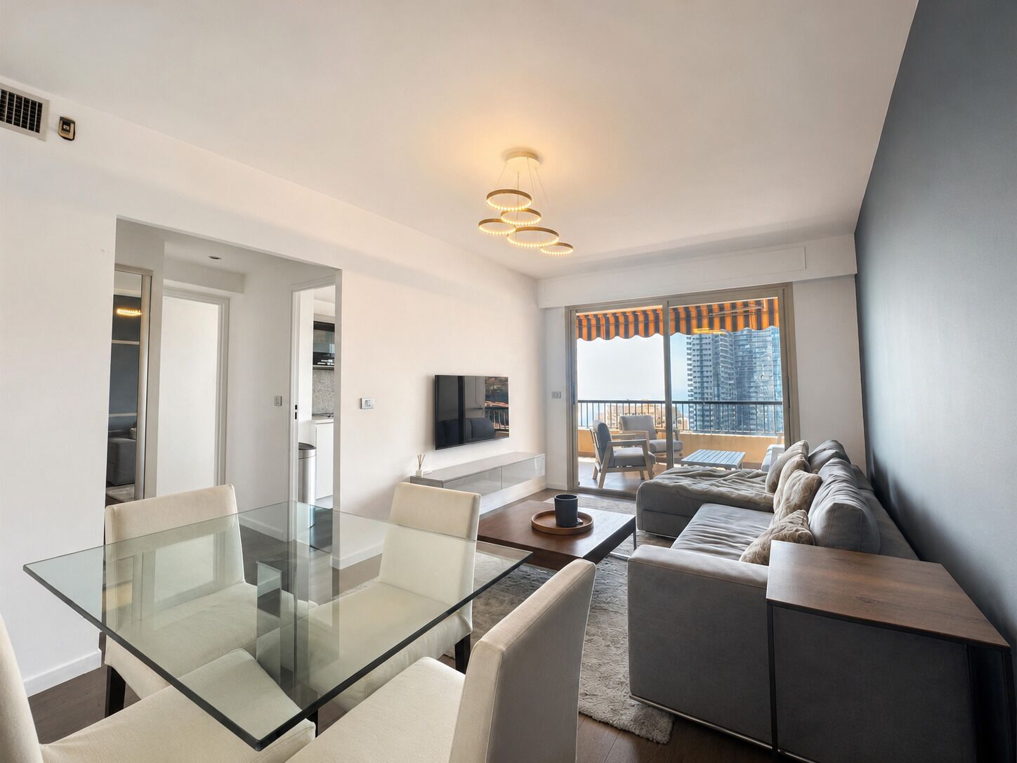 A modern living room with a glass dining table, a sofa, and a TV on the wall.