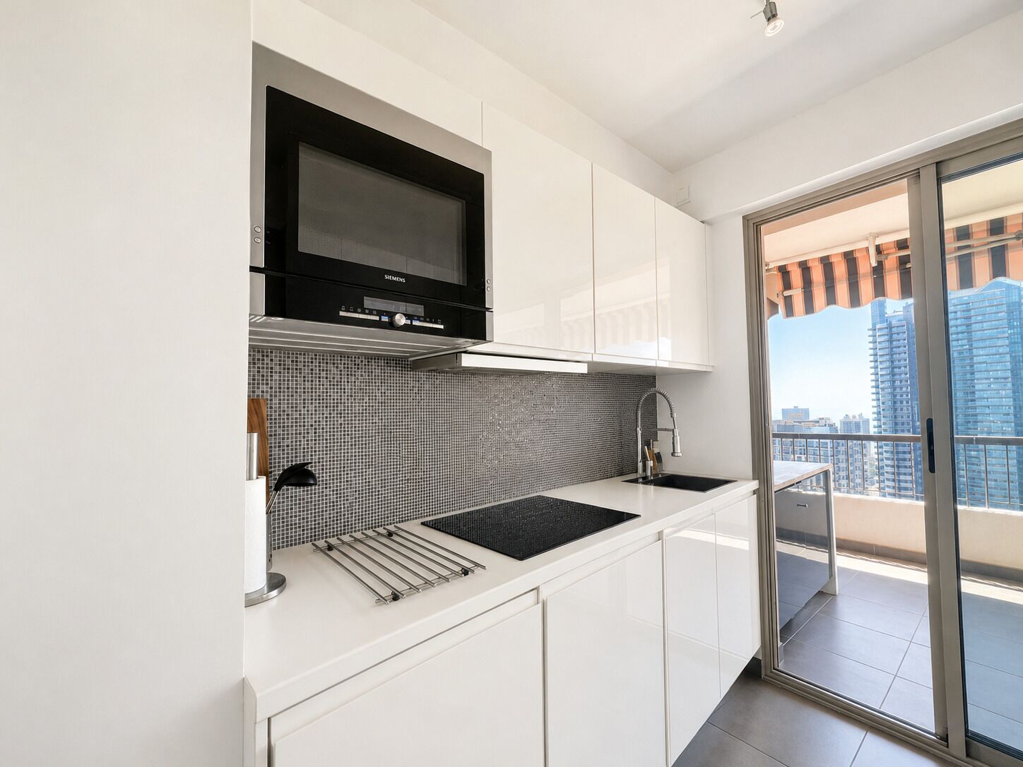 A modern kitchen with a black oven, white cabinets, a tiled backsplash, and a view of a cityscape through a sliding glass door.
