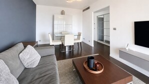 Un salon moderne avec un canapé gris, une table basse avec un plateau, un coin repas avec une table et des chaises blanches, et un téléviseur fixé au mur.