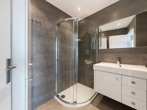 Une salle de bain moderne avec une douche à parois de verre, un meuble-lavabo blanc avec un miroir et des murs recouverts de carreaux foncés.
