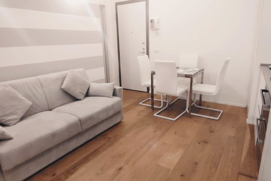 A modern, minimalist living room with a sofa, a dining table with chairs, and a wooden floor.