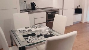 Moderni keittiö, jossa on valkoiset kaapit, lasipintainen ruokapöytä ja musta käsilaukku seinällä.