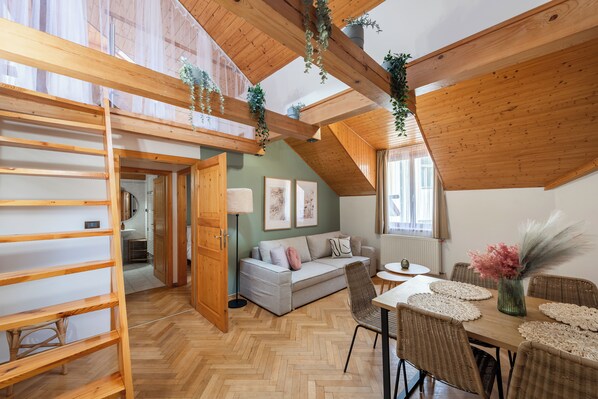 A cozy loft-style room with a wooden ceiling, a herringbone floor, a sofa, a dining table with placemats, and a hanging plant. - Luxury 2BR Apartment Steps from Old Town Square (Praha)