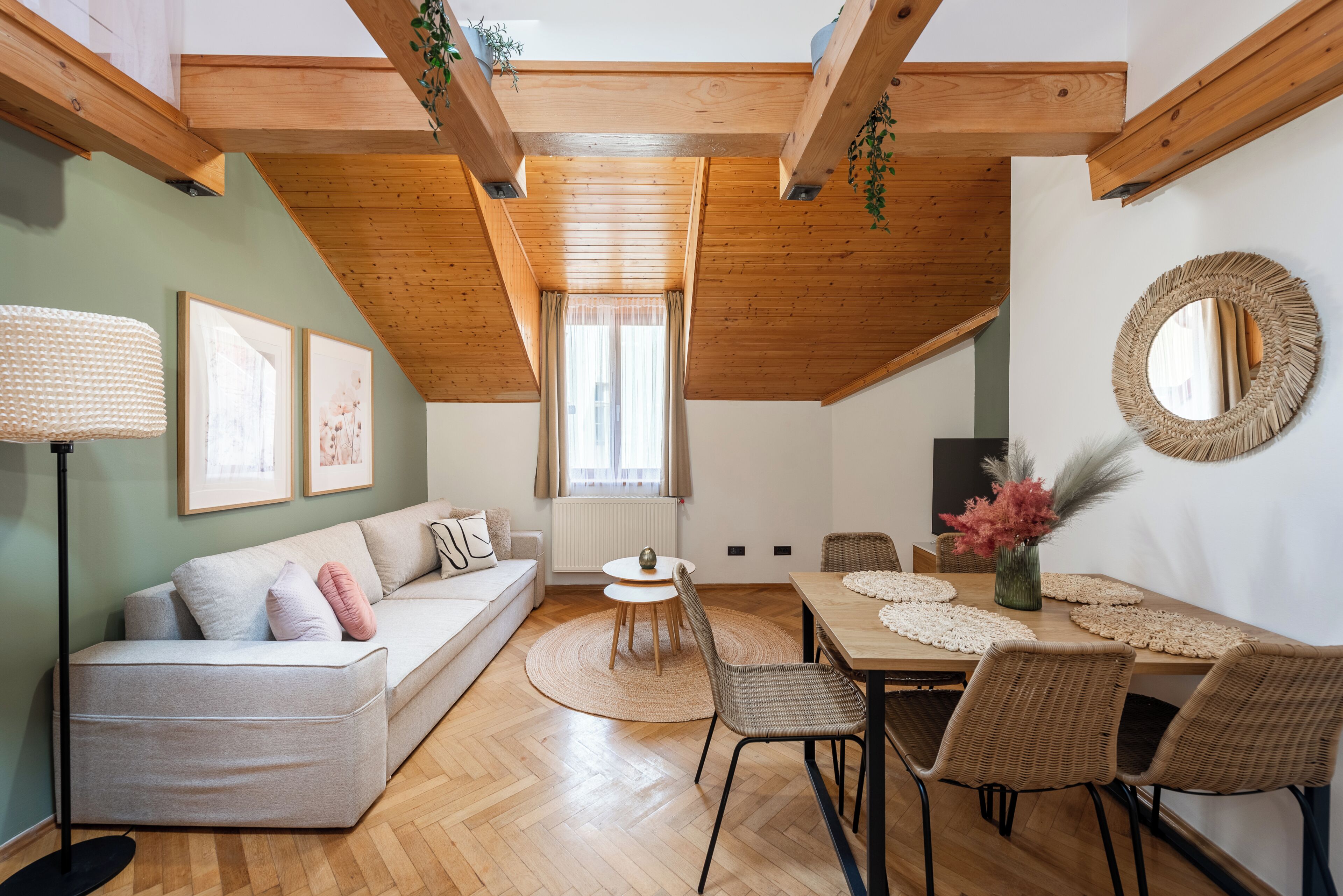 A cozy loft-style room with a high ceiling, wooden beams, a sofa, dining table, chairs, and a large mirror.