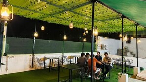An outdoor seating area with tables and chairs under a green canopy, surrounded by a white wall and a green fence.