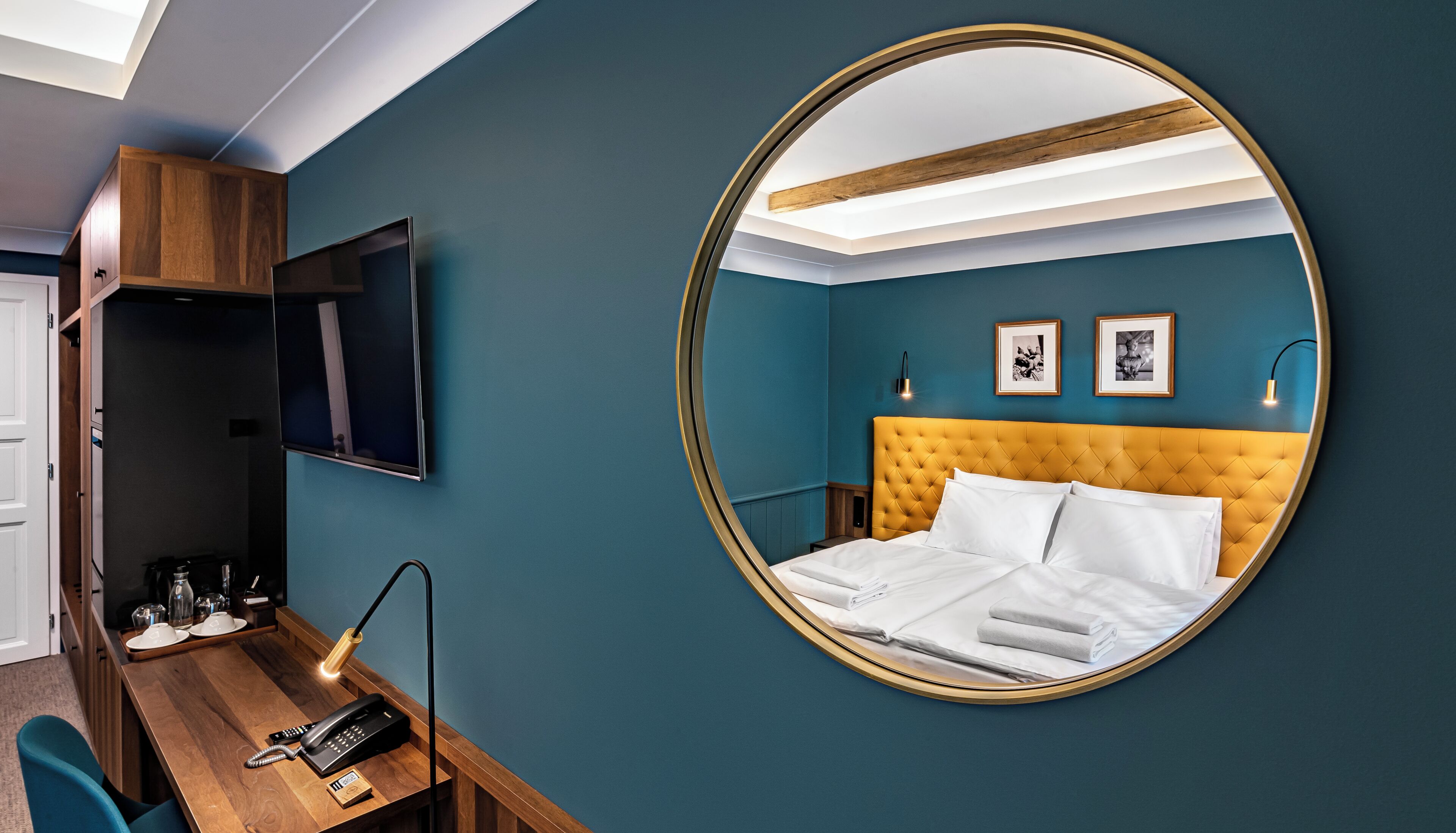 A hotel room with a round mirror, a bed with white linens, a wooden desk with a telephone, and a television mounted on the wall.