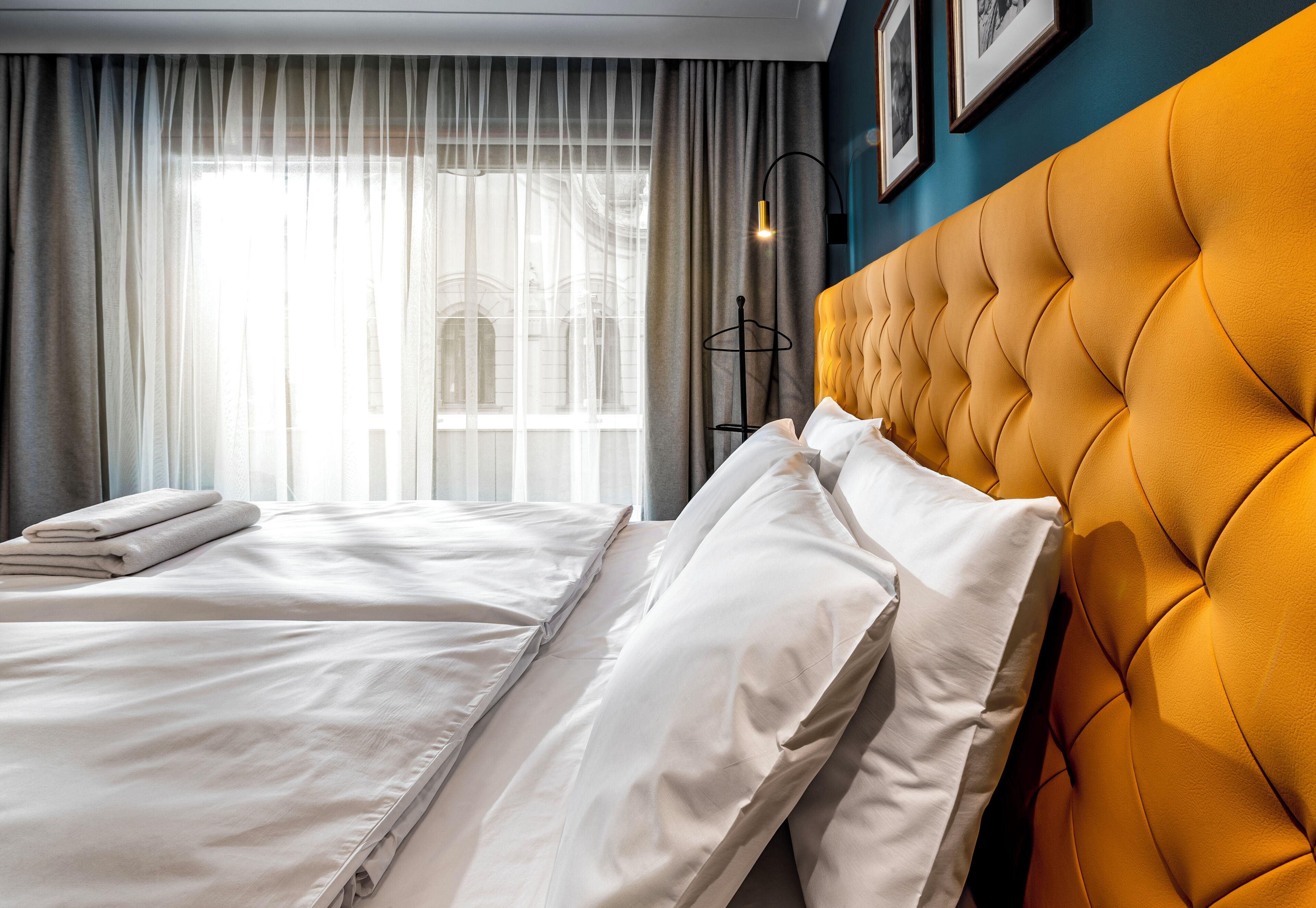 A hotel room with a bed, a headboard, a window with curtains, and a wall-mounted light fixture.