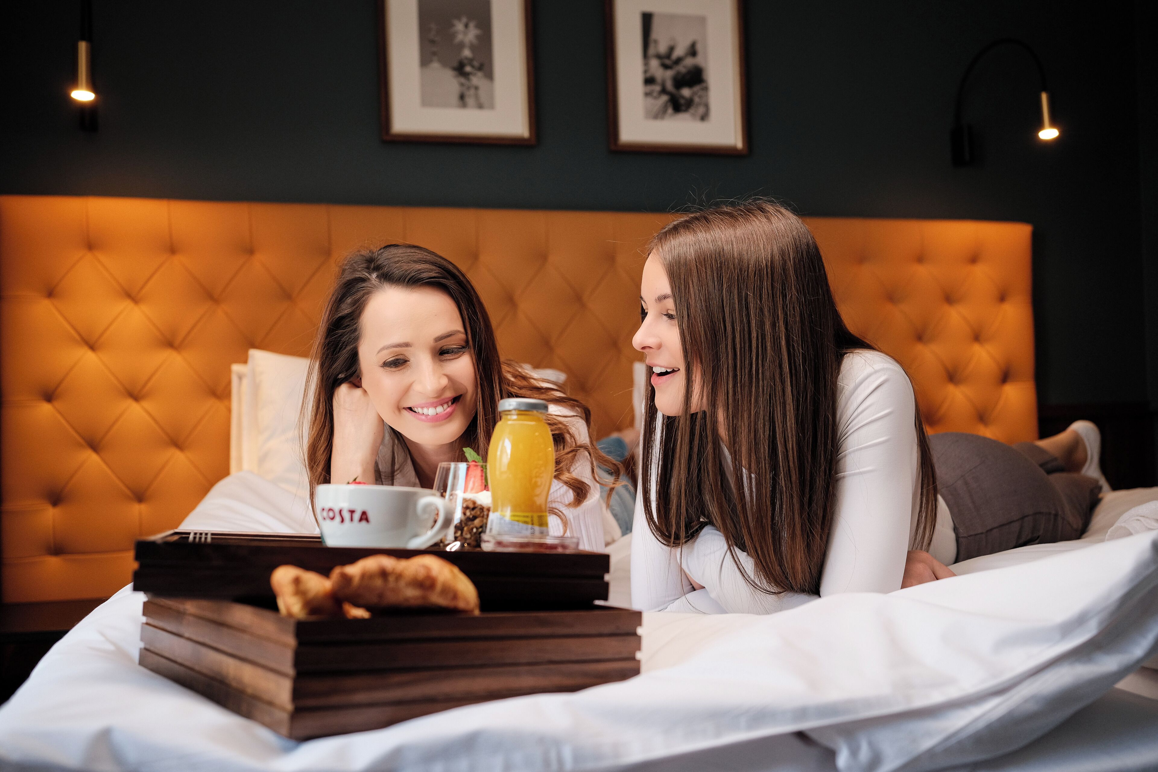 Una habitación con cabecera acolchada, una mesita de noche con una bandeja que contiene una taza, un vaso de jugo y un pastel.