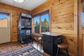 A room with a wooden interior, a gaming machine, a small table, and a window with a view of trees. - New Listing, Game Room, Theater Room, Free Attractions (Sevierville)