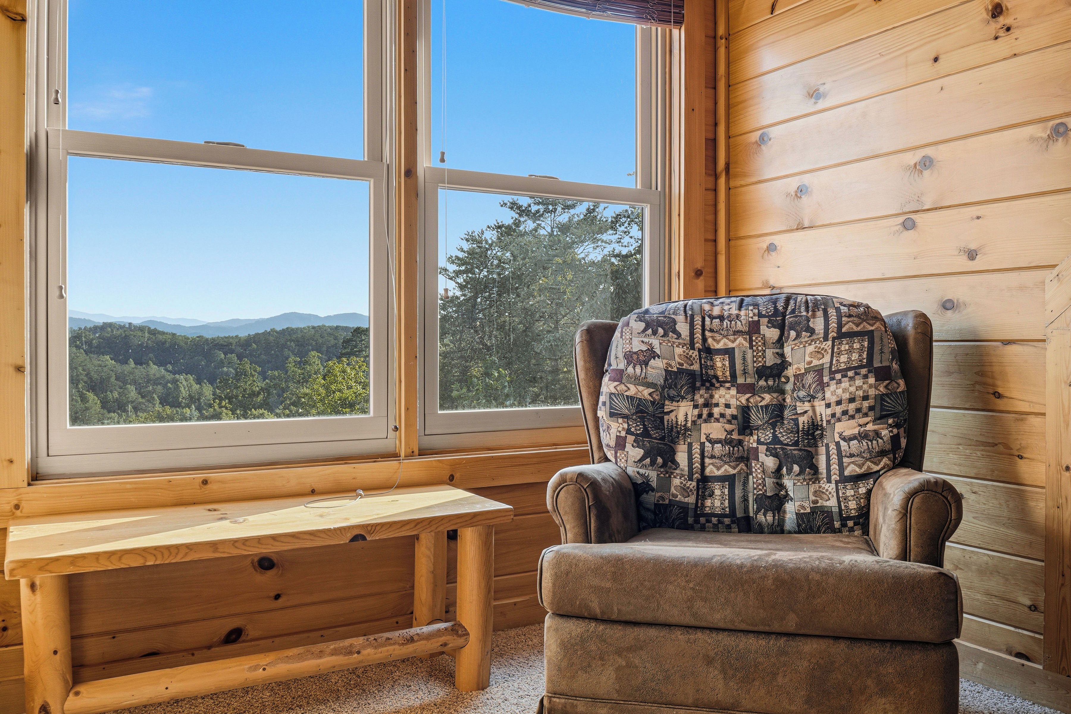 A wooden cabin interior with a large window, a cushioned chair, and a wooden bench.