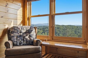 Ein Holzhütten-Innenraum mit einem gepolsterten Sessel, der auf ein großes Fenster mit Blick auf ein bewaldetes Gebirge ausgerichtet ist.