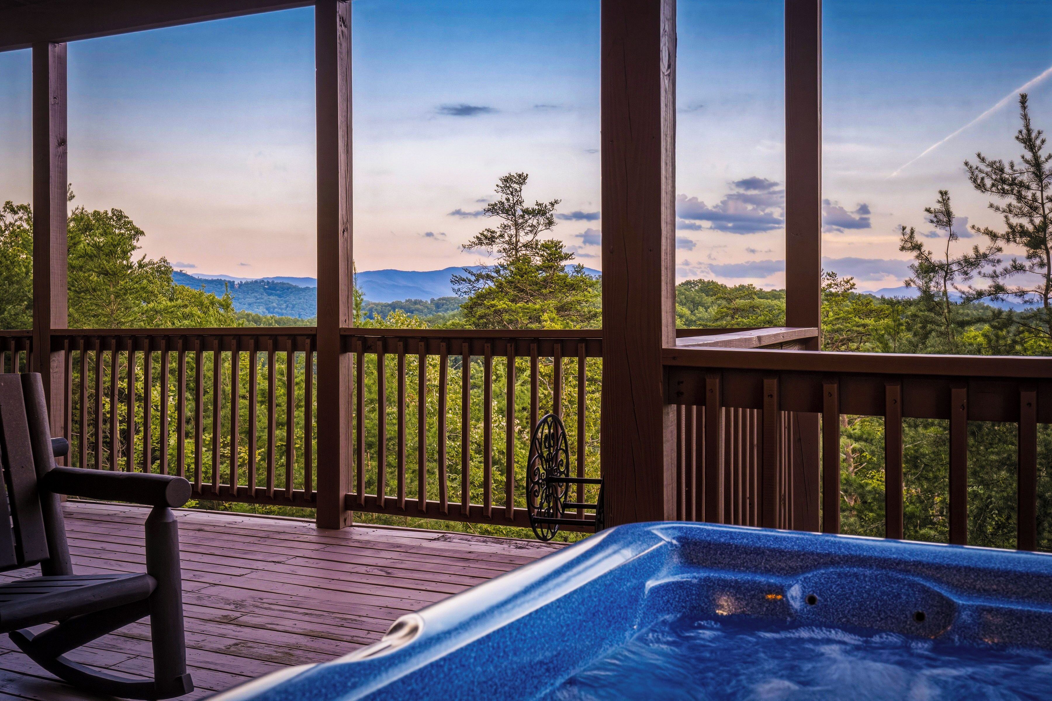 Une terrasse en bois avec un jacuzzi et une chaise, offrant une vue sur un paysage boisé avec des montagnes à l’horizon.