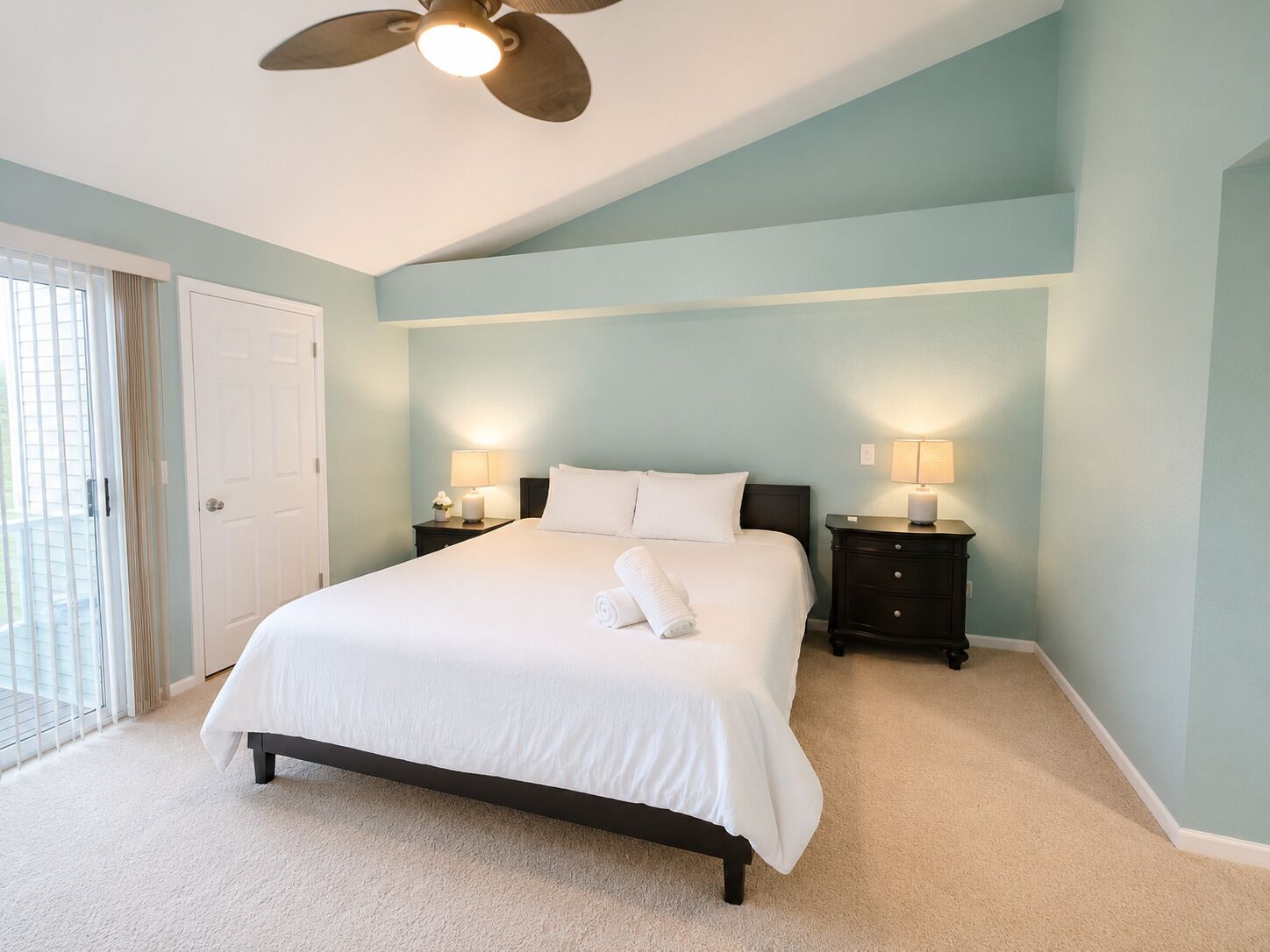 Quarto com cama, duas mesinhas de cabeceira, ventilador de teto e uma porta.