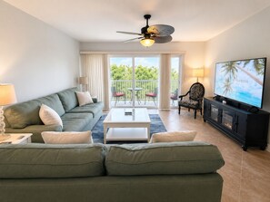 A living room with a sofa, coffee table, TV, and a balcony with chairs. - Luxury Retreat Near Indian Rocks Beach Pool • Game Room • Sleeps 8+ (Seminole)