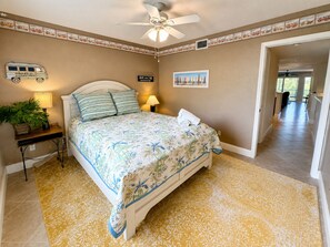 A bedroom with a bed, bedside lamps, a wall-mounted sign, a ceiling fan, and a hallway leading to another room. - Luxury Retreat Near Indian Rocks Beach Pool • Game Room • Sleeps 8+ (Seminole)