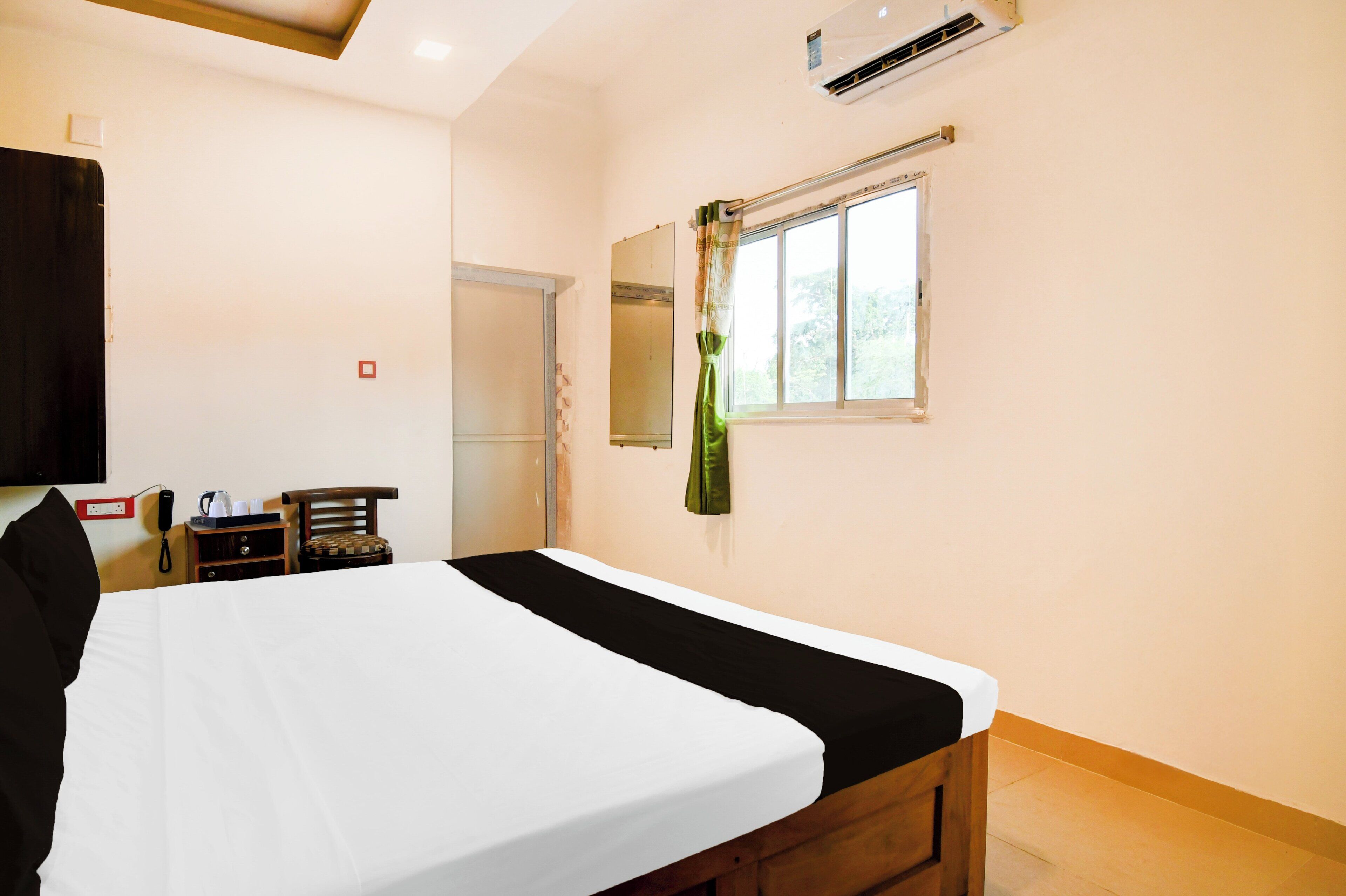 Una habitación de hotel con cama, mesita de noche, una silla, ventana con vistas, unidad de aire acondicionado y teléfono instalado en la pared.