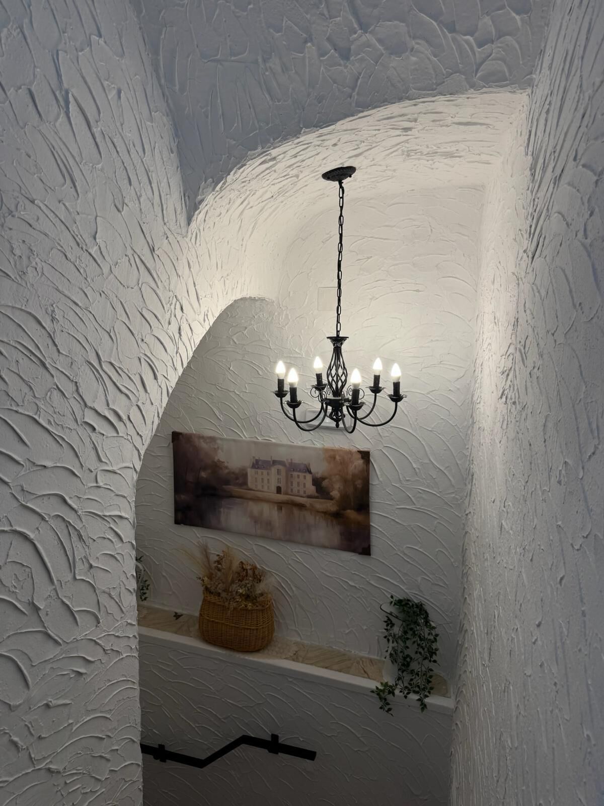 Una habitación con pared blanca texturizada, una lámpara de araña, un cuadro enmarcado y una pequeña planta.