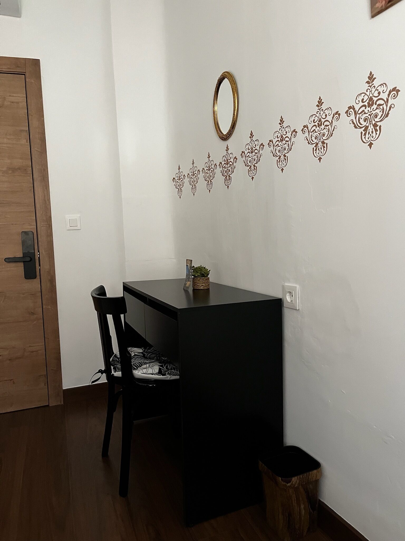 A small room with a wooden door, a black desk, a chair with a patterned cushion, a small plant on the desk, and decorative wall stickers.
