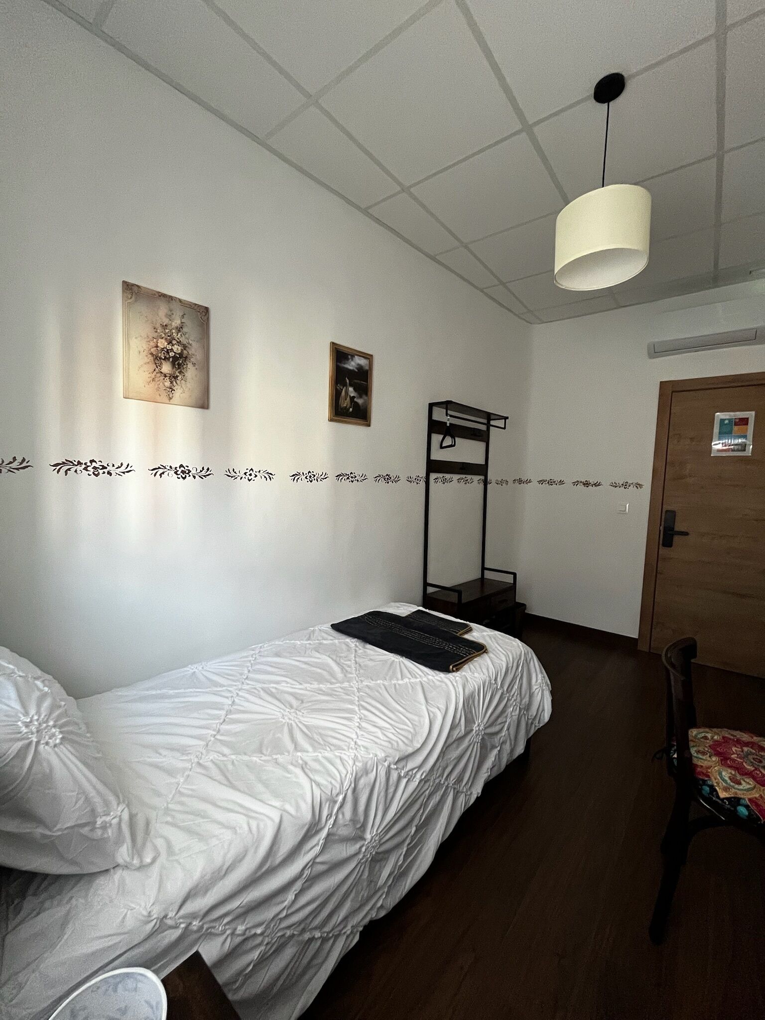 A single-bed room with a bunk bed against the wall, a hanging light fixture, and a door with a sign.