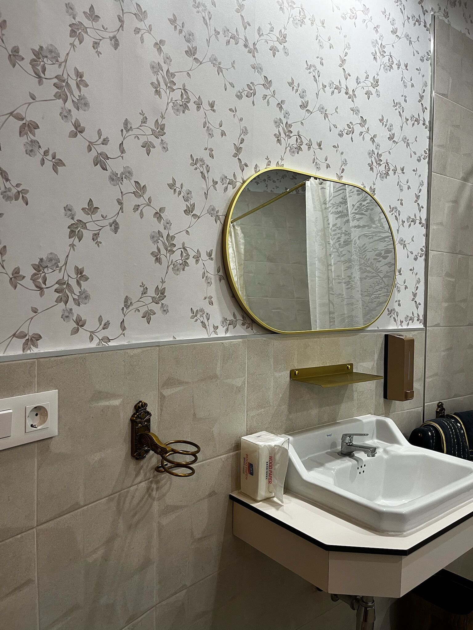 Un baño con un lavamanos blanco, un espejo con marco dorado, papel tapiz floral y un perchero para toallas.