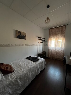 Habitación individual con cama individual y literas, lámpara colgante, un cuadro en la pared y ventana con cortinas.