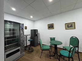 Una habitación con refrigerador, máquina expendedora, mampara de vidrio y una mesa con sillas.