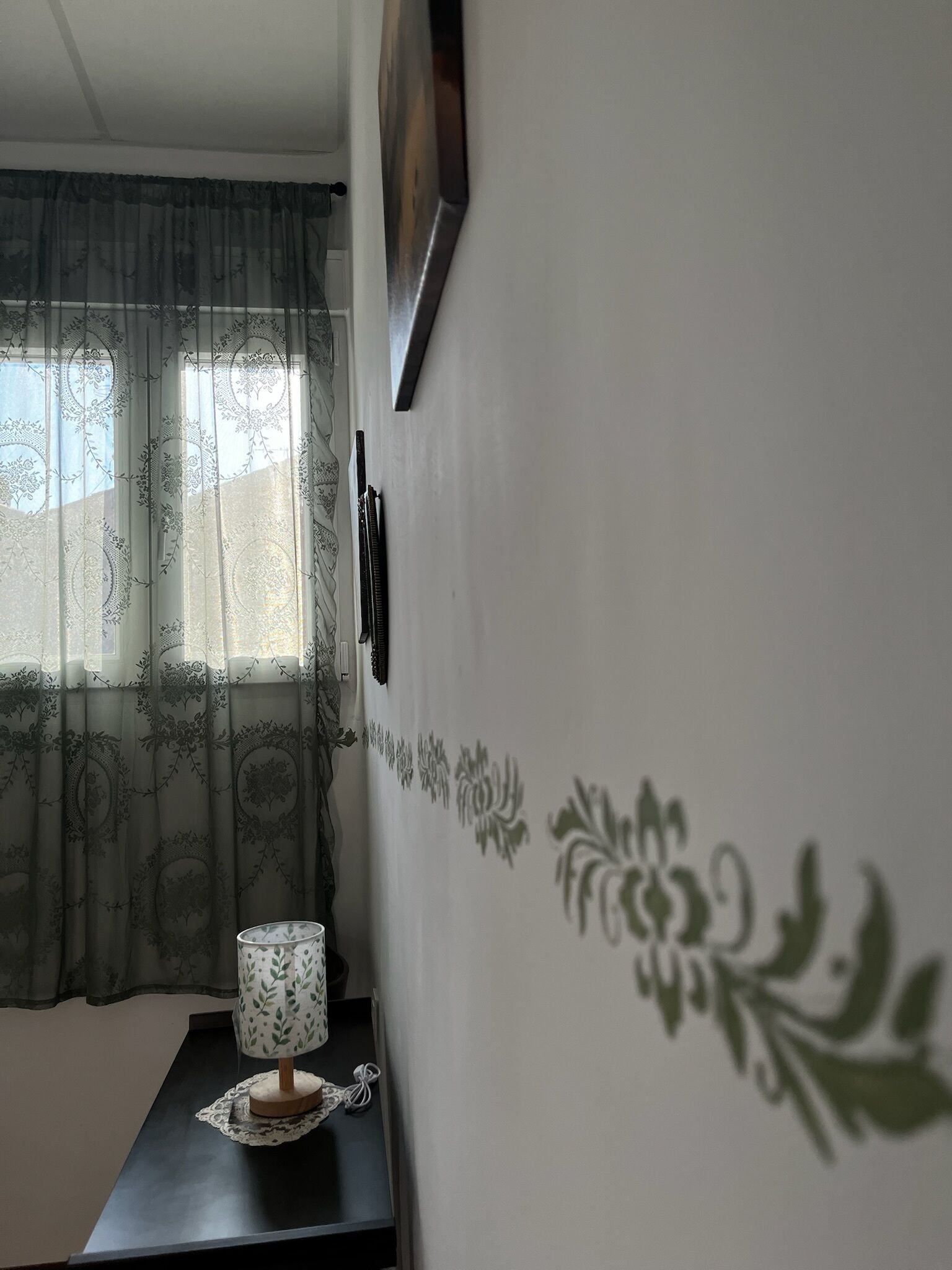 Una habitación con una ventana cubierta por cortinas translúcidas, una pared con motivos florales y una mesa con una lámpara.