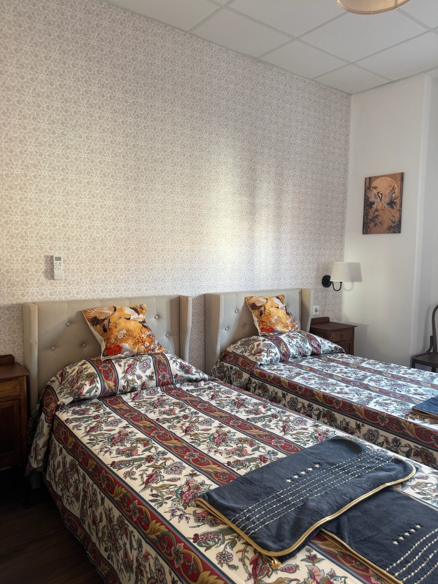 En dobbeltseng med blomstret sengetøj og to puder, et natbord, en vægmonteret lampe og et billede på væggen.