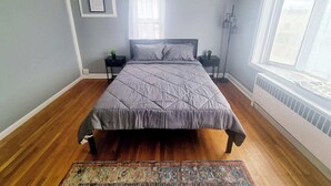 A bedroom with a bed, two bedside tables, a floor lamp, a window with blinds, and a radiator. - Inviting 1BR Apartment — Rockledge Location (Rockledge)