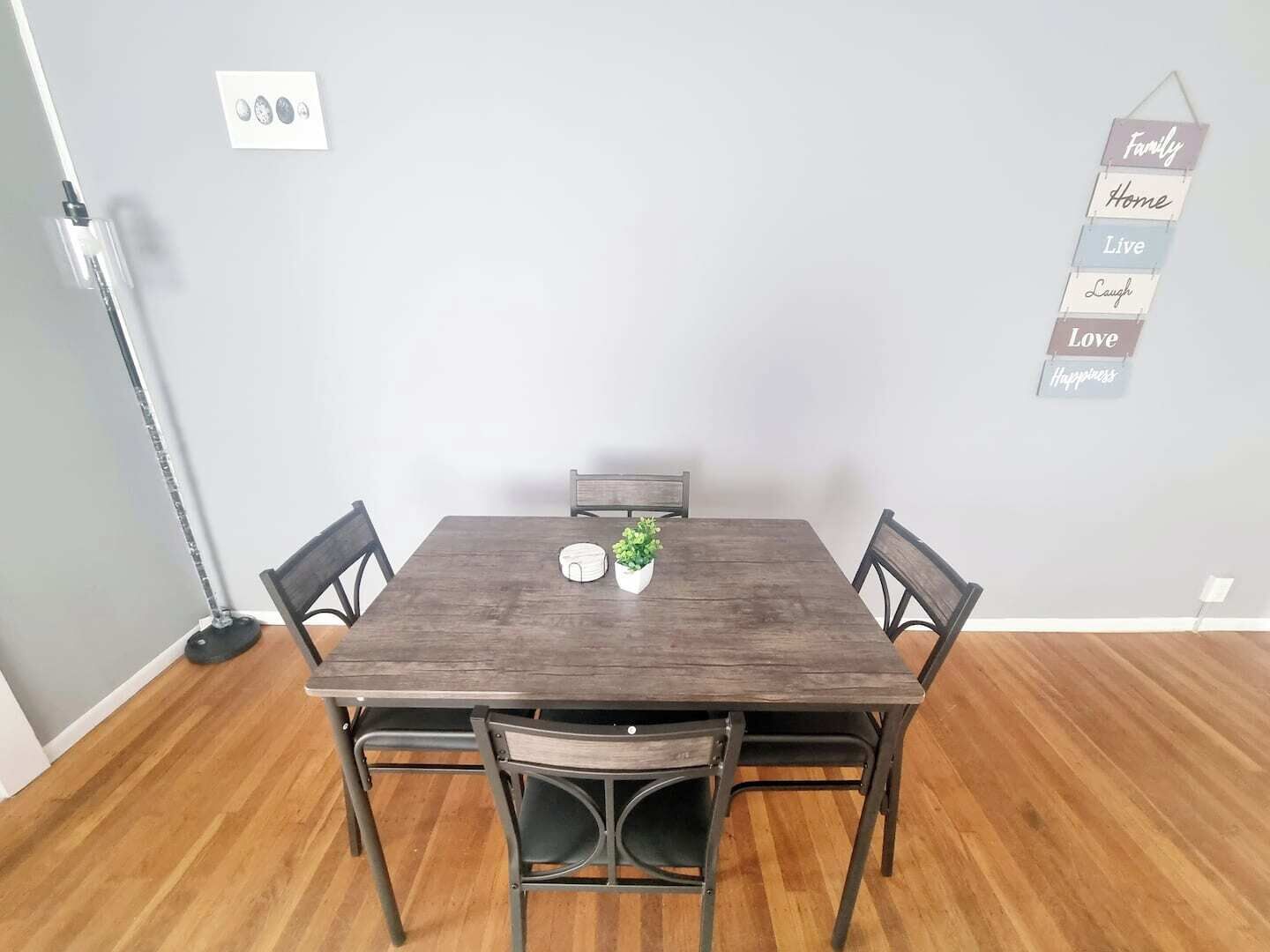 A dining area with a wooden table and four chairs, a small potted plant, and a wall-mounted sign with positive phrases.