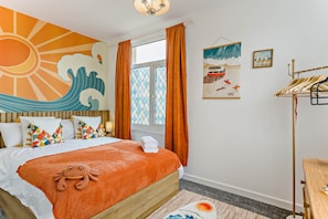 Una habitación de hotel con cama, ventana con cortinas, un cuadro de una ola en la pared y suelo de madera.