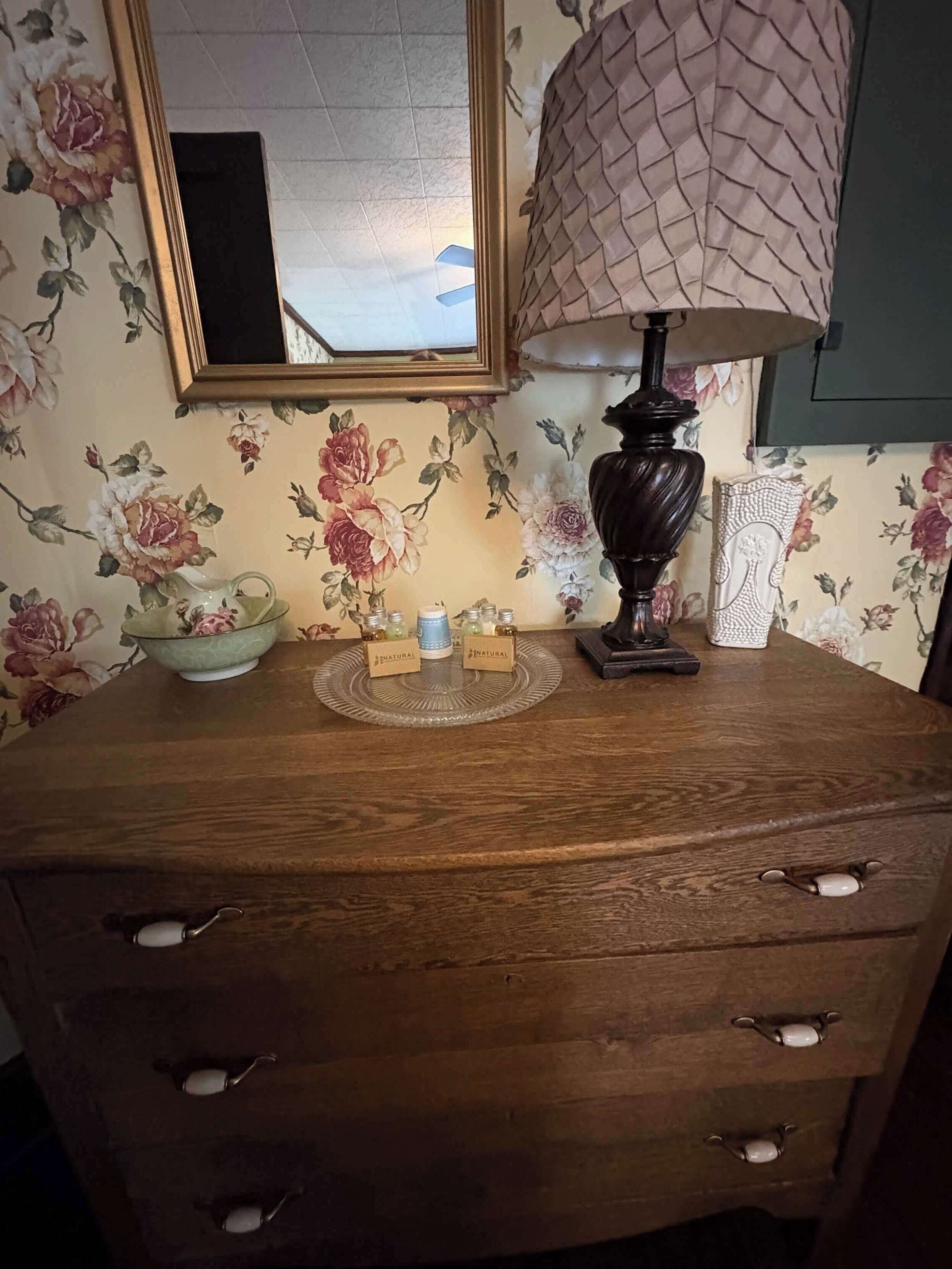 A wooden dresser with three drawers, a lamp, and a floral wallpaper.