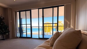 A room with a large sliding glass door leading to a balcony with a view of the sea and autumn trees. - Bluewater Sanctuary: Luxury Beach house with private jetty (Tascott)