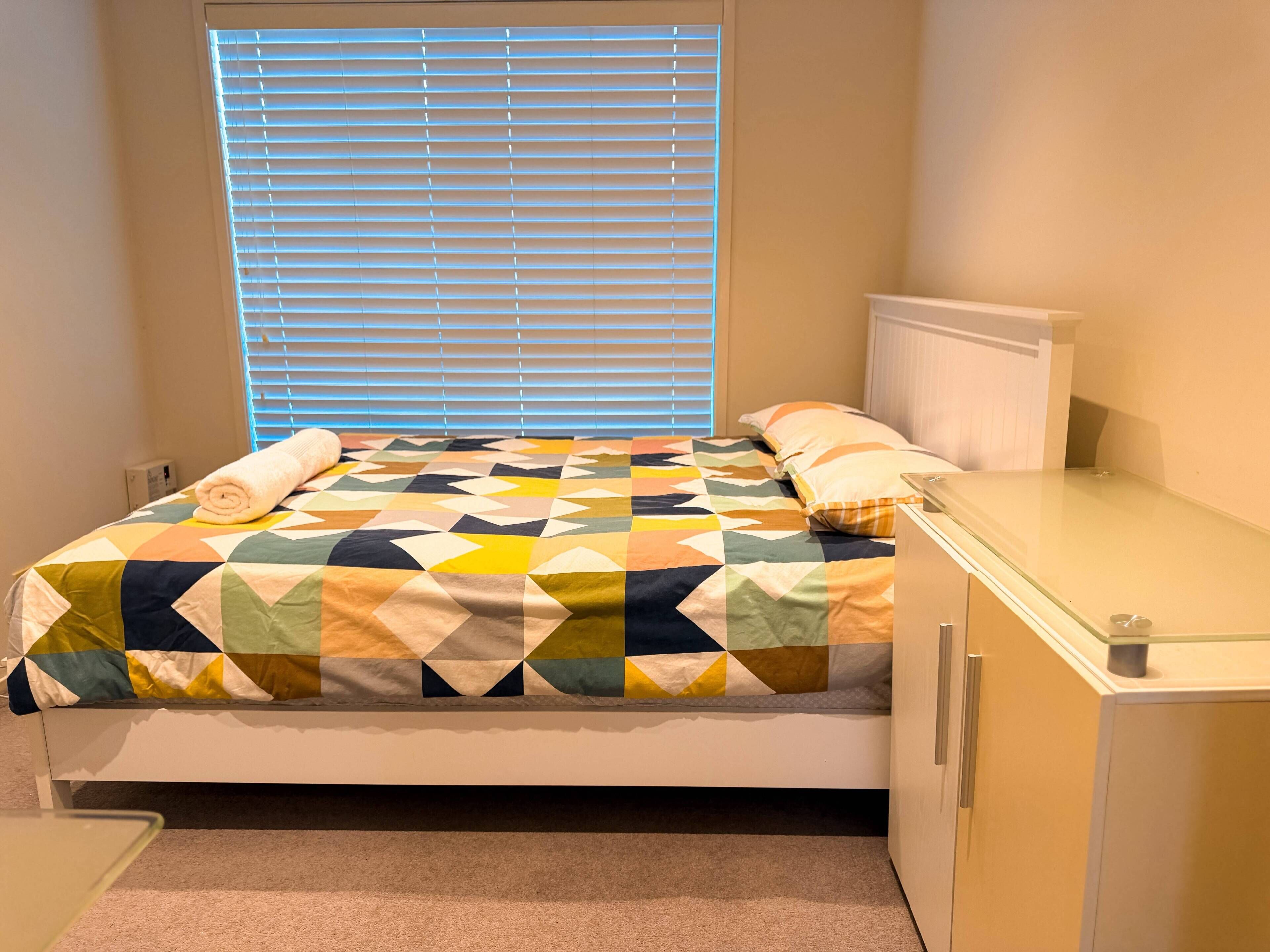 Ein Bett mit einer quiltartigen Steppdecke mit geometrischem Muster, ein Nachttisch und ein Fenster mit Jalousien.
