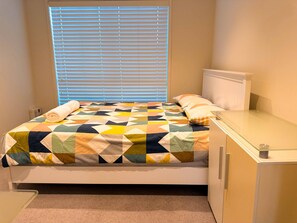 A bed with a geometric-patterned quilt, a bedside table, and a window with blinds. - Bluewater Sanctuary: Luxury Beach house with private jetty (Tascott)