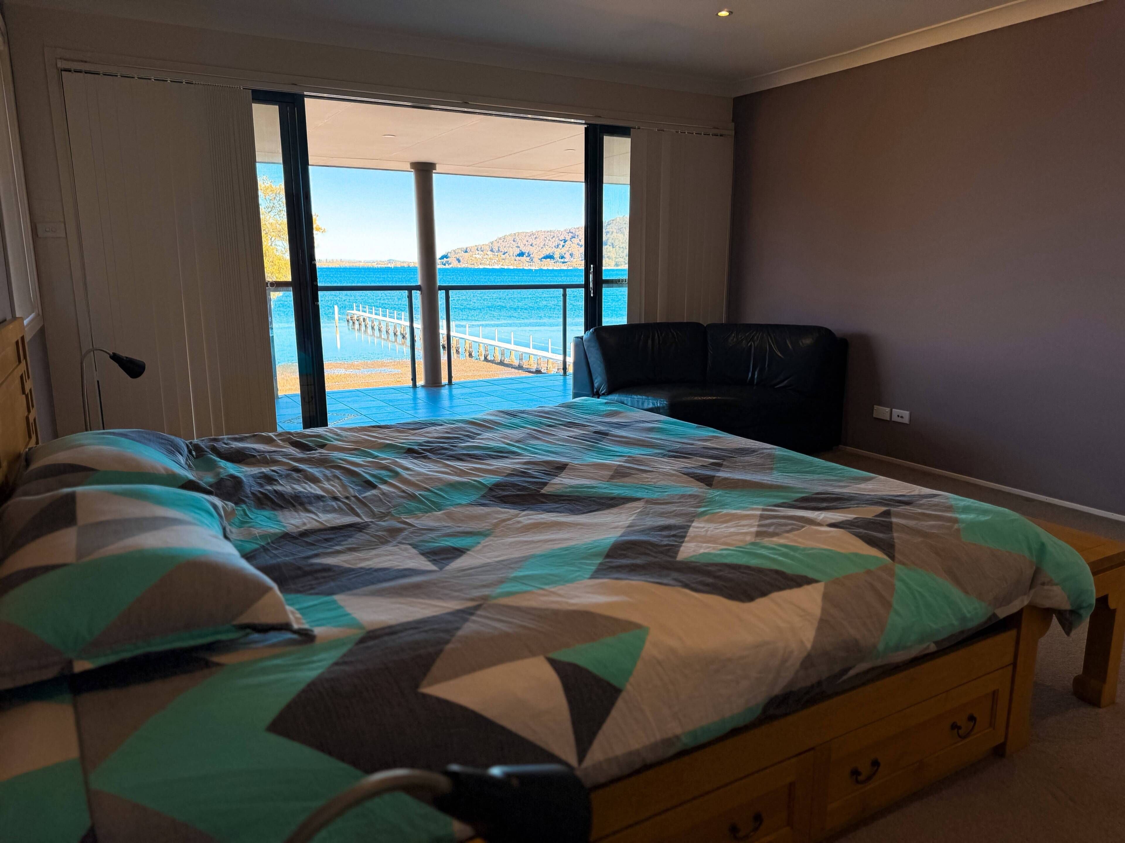 A bedroom with a bed, a wooden nightstand, and a view of a beach and pier through a large window.